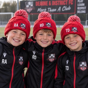 May include: Three red knit beanies with white pom-poms, each featuring a rectangular patch with the Dearne & District FC logo and initials RA, RD, or TD. The beanies are worn with matching black and red athletic zip-up jackets with club logos.