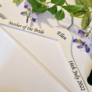 May include: A white wooden hanger with the text "Mother of the Bride", the name "Ellen", and the date "16th July 2022" in black script. The hanger is decorated with purple flowers and green leaves.