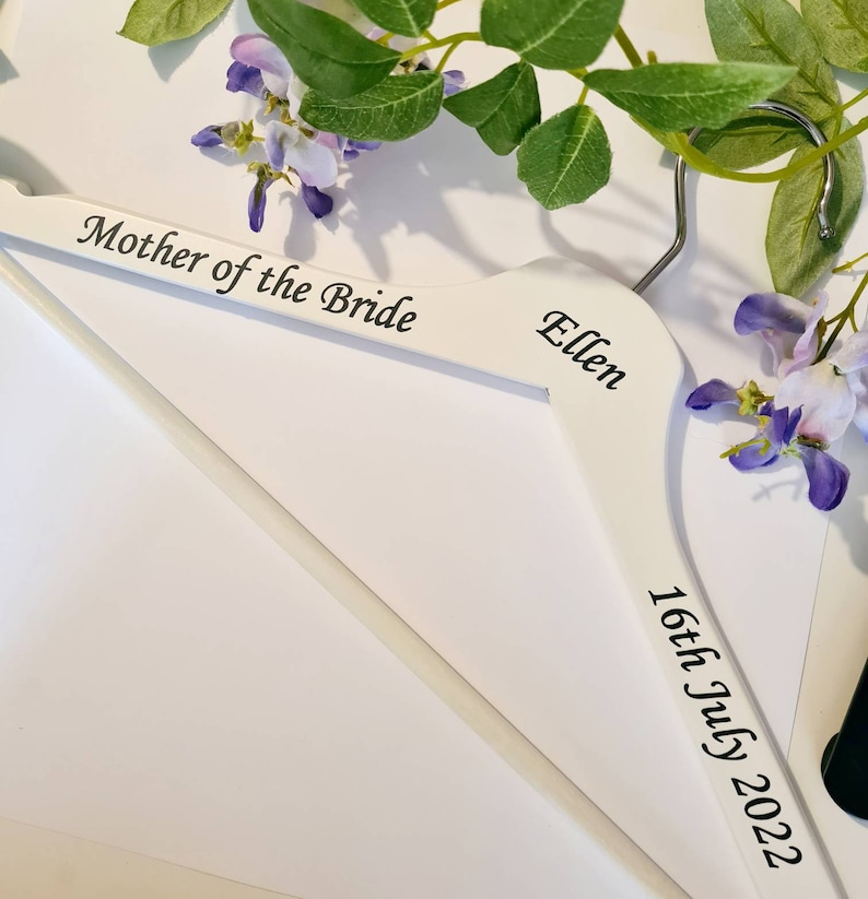 May include: A white wooden hanger with the text "Mother of the Bride", the name "Ellen", and the date "16th July 2022" in black script. The hanger is decorated with purple flowers and green leaves.