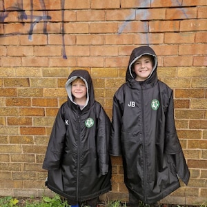 Puede incluir: Dos niños con chaquetas negras con capucha, logotipos verdes e iniciales blancas. Las chaquetas tienen cierre de cremallera y se llevan sobre pantalones negros. Los niños están de pie frente a una pared de ladrillo con grafitis.