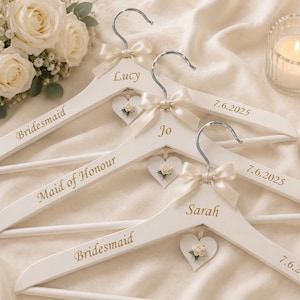May include: Three white wooden clothes hangers featuring silver metal hooks, cream-colored satin bows, and small heart-shaped tags with floral accents. Each hanger is personalized with gold text reading Lucy, Jo, Sarah, Bridesmaid, Maid of Honour, and 7.6.2025.