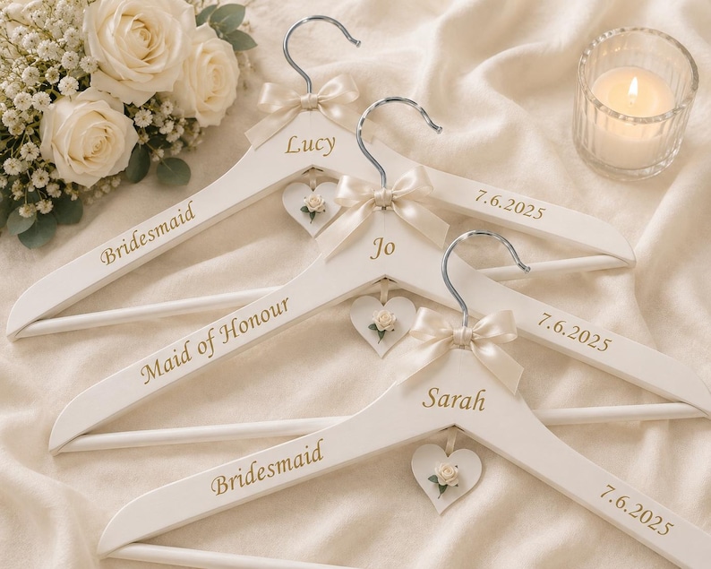 May include: Three white wooden clothes hangers featuring silver metal hooks, cream-colored satin bows, and small heart-shaped tags with floral accents. Each hanger is personalized with gold text reading Lucy, Jo, Sarah, Bridesmaid, Maid of Honour, and 7.6.2025.