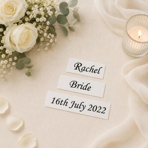 May include: Three rectangular white paper labels with black printed text. The top label reads Rachel, the middle label reads Bride, and the bottom label reads 16th July 2022. The labels are arranged vertically on a light-colored fabric surface.