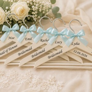 May include: Five white wooden clothes hangers with silver metal hooks, each adorned with a light blue satin bow. The hangers are personalized with black text including names like Mae, Emil, Rachel, Lucy, Lydia, and titles such as Bride and Bridesmaid.