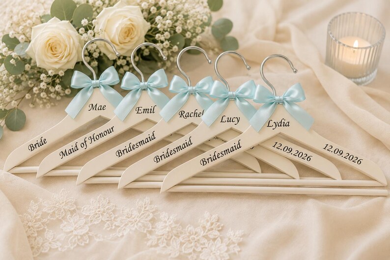 May include: Five white wooden clothes hangers with silver metal hooks, each adorned with a light blue satin bow. The hangers are personalized with black text including names like Mae, Emil, Rachel, Lucy, Lydia, and titles such as Bride and Bridesmaid.