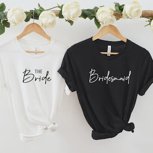 Könnte beinhalten: Zwei T-Shirts, eines weiß und eines schwarz, hängen an Holzbügeln. Auf dem weißen Shirt steht "The Bride" in Schreibschrift, und auf dem schwarzen Shirt steht "Bridesmaid". Weiße Rosen und Grünzeug schmücken den oberen Teil des Bildes. Hochzeitskleidung.