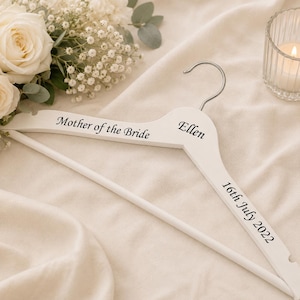 May include: A white wooden clothes hanger featuring a silver metal hook and black printed text that reads Mother of the Bride, the name Ellen, and the date 16th July 2022, positioned along the top and side edges of the hanger frame.