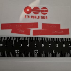 Può includere: Un foglio di adesivi trasparente con il logo rosso BTS WORLD TOUR e barre rettangolari rosse con i nomi RM, JIN, SUGA, J-HOPE, JIMIN e JUNG KOOK scritti in bianco. Un righello nero posizionato sotto indica una larghezza di 10 centimetri.
