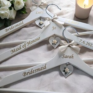May include: White wooden hangers with silver hooks, personalized with names and titles in gold lettering. Each hanger has a cream-colored bow and a heart-shaped charm with a small flower. The hangers are for a wedding.
