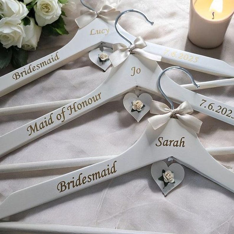 May include: White wooden hangers with silver hooks, personalized with names and titles in gold lettering. Each hanger has a cream-colored bow and a heart-shaped charm with a small flower. The hangers are for a wedding.