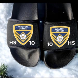Puede incluir: Un par de sandalias negras con el logotipo de Walton United en amarillo, azul y blanco. Las sandalias muestran el número "10" y las letras "HS". El calzado se presenta sobre un fondo nevado, con un toque de vegetación.