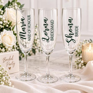 May include: Three clear glass champagne flutes with stems. The first glass features the text Maria and Maid of Honour, the second Lorna and Bridesmaid, and the third Shona and Bride in dark green lettering. A small box nearby reads Bridal Party.