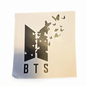 K&ouml;nnte beinhalten: Ein wei&szlig;er Aufkleber mit den Buchstaben "BTS" in fetter, serifenloser Schrift. Das Design beinhaltet ein stilisiertes Emblem mit Schmetterlingen, die von oben herauskommen und einen dynamischen visuellen Effekt erzeugen. Die Schmetterlinge sind in verschiedenen Gr&ouml;&szlig;en.