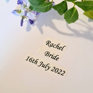 May include: Three white rectangular stickers with black text. The top sticker reads "Rachel", the middle sticker reads "Bride", and the bottom sticker reads "16th July 2022". The stickers are on a white surface, with purple and green floral accents.