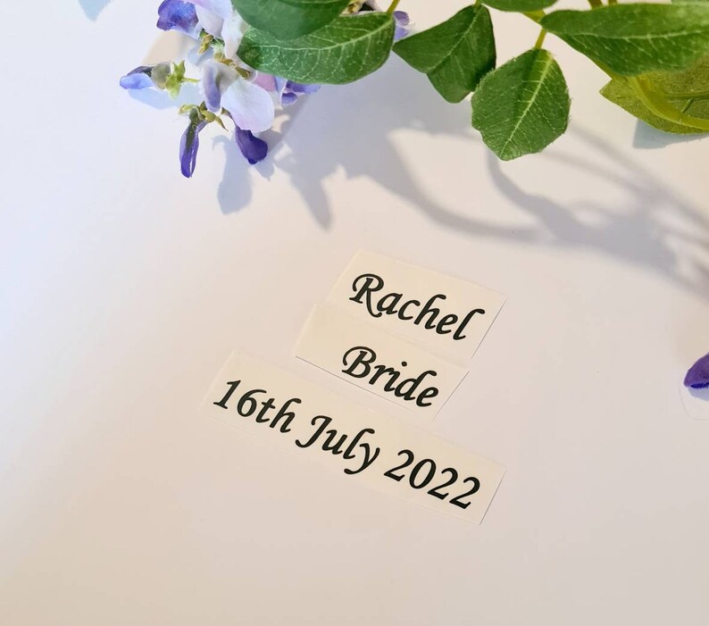May include: Three white rectangular stickers with black text. The top sticker reads "Rachel", the middle sticker reads "Bride", and the bottom sticker reads "16th July 2022". The stickers are on a white surface, with purple and green floral accents.