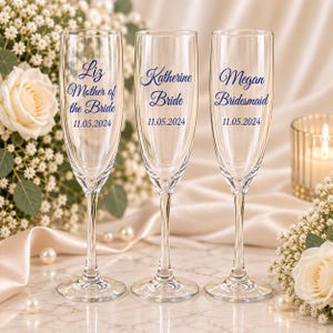 May include: Three clear glass champagne flutes with blue script text. The first reads Liz Mother of the Bride 11.05.2024, the second Katherine Bride 11.05.2024, and the third Megan Bridesmaid 11.05.2024. Each glass has a tall, slender bowl.