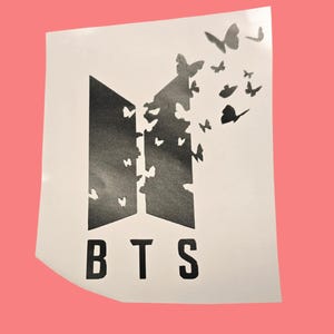 Puede incluir: Una pegatina blanca con las letras "BTS" en negrita, en negro. El diseño incorpora un logotipo negro estilizado con mariposas, algunas de las cuales parecen estar rompiendo el logotipo. El fondo es de color coral claro.