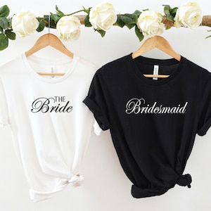 Op de afbeelding: Twee T-shirts hangen aan houten hangers. Het witte T-shirt heeft de tekst "The Bride" in zwarte schrijfletters. Het zwarte T-shirt heeft de tekst "Bridesmaid" in witte schrijfletters.