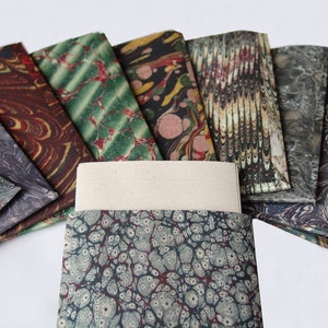 Peut inclure: Un ensemble de feuilles de papier marbr&eacute; color&eacute; dans diff&eacute;rents motifs, y compris le vert, le rouge, le brun et le noir. Les feuilles sont dispos&eacute;es en &eacute;ventail, avec une feuille blanche vierge au centre.