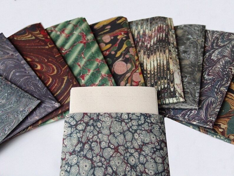 Peut inclure: Un ensemble de feuilles de papier marbr&eacute; color&eacute; dans diff&eacute;rents motifs, y compris le vert, le rouge, le brun et le noir. Les feuilles sont dispos&eacute;es en &eacute;ventail, avec une feuille blanche vierge au centre.