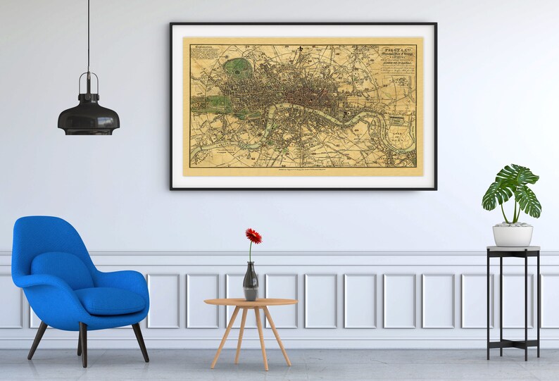 Peut inclure: Une carte vintage de Londres, en Angleterre, encadr&eacute;e en noir, est accroch&eacute;e &agrave; un mur blanc au-dessus d'un fauteuil bleu et d'une petite table en bois avec un vase de fleurs rouges. Une plante en pot est plac&eacute;e sur un support en m&eacute;tal noir &agrave; droite.
