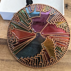 May include: A circular, copper-colored ornament with a tree-like design. The ornament is filled with colorful, translucent resin, creating a stained glass effect. The design is reminiscent of a phylogenetic tree, representing the evolutionary relationships between different species.