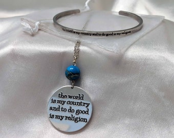 May include: A silver necklace with a round pendant that says "the world is my country and to do good is my religion". The pendant is attached to a blue gemstone. The necklace is on a white satin background with a silver bangle bracelet that says "the world is my country and to do good is my religion".
