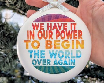 May include: White round Christmas ornament with the text "WE HAVE IT IN OUR POWER TO BEGIN THE WORLD OVER AGAIN" in colorful letters. The ornament has a white ribbon for hanging.