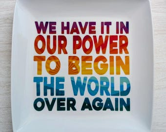 May include: A square white ceramic plate with rounded corners. The plate features the quote, "WE HAVE IT IN OUR POWER TO BEGIN THE WORLD OVER AGAIN," in a gradient of colors from purple to blue.