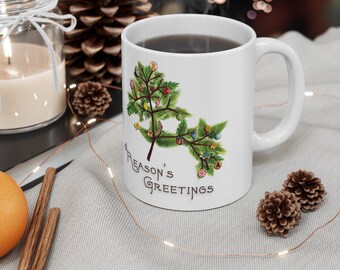 May include: White ceramic mug with a green Christmas tree design and the text "Reason's Greetings". The mug is filled with coffee and steam is rising from the top.