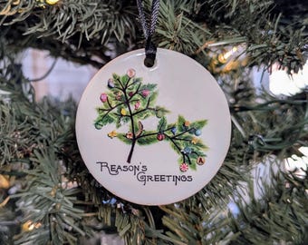 May include: A round, off-white Christmas ornament with a painted Christmas tree design. The tree is decorated with colorful ornaments. The words "Reason's Greetings" are written in black script. A black ribbon is attached for hanging.