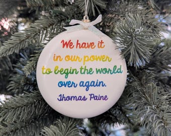 May include: White, round Christmas ornament with the quote "We have it in our power to begin the world over again. Thomas Paine" in rainbow colors. The ornament hangs from a silver string and bow, set against a backdrop of a green Christmas tree.