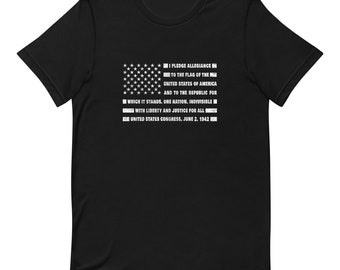 May include: Black t-shirt with a white American flag and the Pledge of Allegiance text printed on it. The text reads: "I pledge allegiance to the flag of the United States of America and to the republic for which it stands, one nation, indivisible, with liberty and justice for all. United States Congress, June 2, 1942."