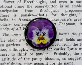 May include: A round, black-rimmed button featuring a close-up of a purple, white, and yellow pansy flower. The button is set against a backdrop of printed text from a book, with visible words like "pansies" and "Hamlet".