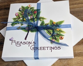 May include: A stack of white greeting cards with a blue ribbon tied around them. The front of the card features a watercolor illustration of a Christmas tree with ornaments and the text "Reason's Greetings".
