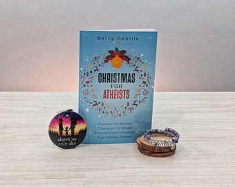 May include: A blue book with the title "Christmas for Atheists" by Betsy DeVille. The book cover features a wreath of Christmas-themed illustrations. A colorful round ornament with a family silhouette and the text "above us only sky" is in the foreground. A purple and silver beaded bracelet is in the lower right corner.