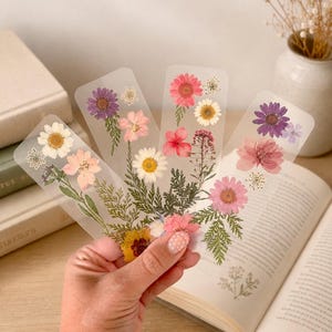 May include: Four rectangular, transparent bookmarks featuring arrangements of pressed, dried flowers and green foliage. The floral designs include pink, purple, white, and yellow blossoms, with delicate stems and leaves embedded within the clear material.