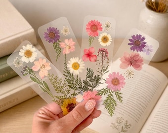 Puede incluir: Cuatro marcapáginas rectangulares transparentes con arreglos de flores prensadas, secas y follaje verde. Los diseños florales incluyen flores rosas, moradas, blancas y amarillas, con tallos y hojas delicadas incrustadas en el material.