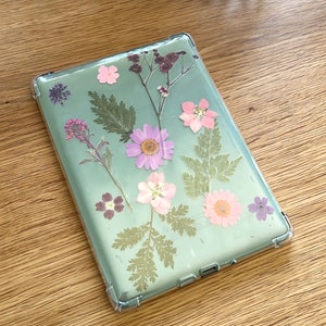 May include: Clear phone case with pressed flowers. The case features a variety of dried flowers in shades of pink, purple, and green, including daisies and ferns. The case is on a wooden surface.
