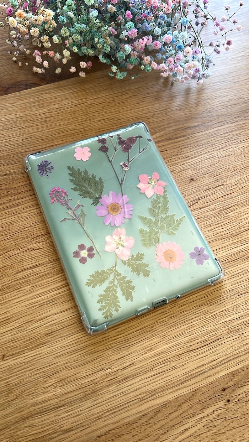 May include: Clear phone case with pressed flowers. The case features a variety of dried flowers in shades of pink, purple, and green, including daisies and ferns. The case is on a wooden surface.
