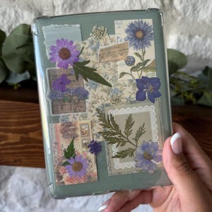 May include: A clear, rectangular case with a green backing, decorated with pressed flowers and botanical ephemera. The design includes purple and blue flowers, fern fronds, and vintage paper elements. The case is held in a hand.