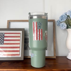Op de afbeelding: Een saliegroene roestvrijstalen beker met een doorzichtig deksel en een doorzichtig rietje. De beker is voorzien van een verticale sticker met een Amerikaans vlagontwerp bestaande uit bloemmotieven in rood, wit en blauw.