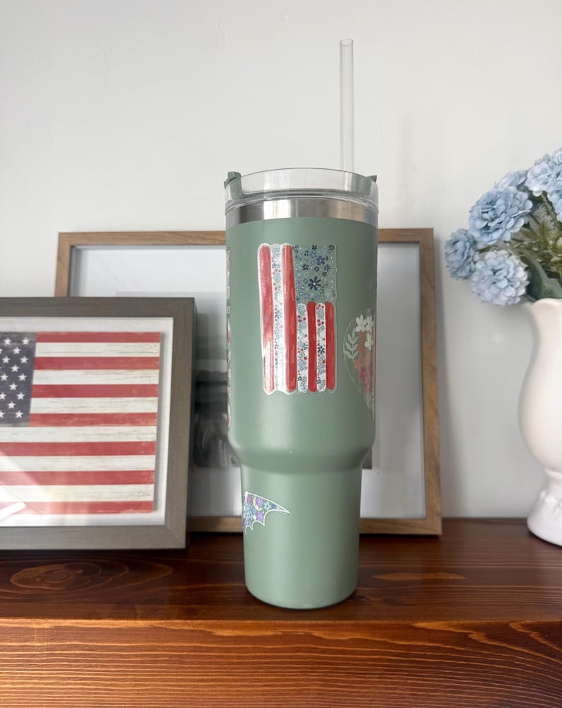 Op de afbeelding: Een saliegroene roestvrijstalen beker met een doorzichtig deksel en een doorzichtig rietje. De beker is voorzien van een verticale sticker met een Amerikaans vlagontwerp bestaande uit bloemmotieven in rood, wit en blauw.