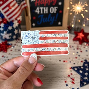 Op de afbeelding: Een rechthoekige sticker met een gestileerd Amerikaans vlagontwerp. Het kanton is gevuld met kleine blauwe en witte bloemen, terwijl de strepen afwisselen tussen effen rode banen en banen versierd met kleine rode, witte en blauwe bloemmotieven.