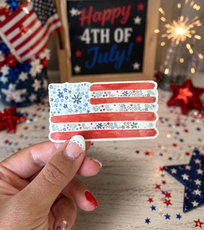 Op de afbeelding: Een rechthoekige sticker met een gestileerd Amerikaans vlagontwerp. Het kanton is gevuld met kleine blauwe en witte bloemen, terwijl de strepen afwisselen tussen effen rode banen en banen versierd met kleine rode, witte en blauwe bloemmotieven.