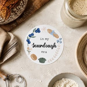 May include: A round sign with the text "in my sourdough era" surrounded by illustrations of blueberries, bread, and baking tools. A loaf of bread, a jar, and a bowl of flour are also visible.