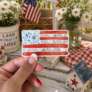 Op de afbeelding: Een rechthoekige sticker met een gestileerd Amerikaans vlagontwerp. De vlag bestaat uit afwisselende horizontale banen van effen rood en wit, gevuld met kleine blauwe, witte en rode bloemmotieven. Het kanton is blauw met bloemen.