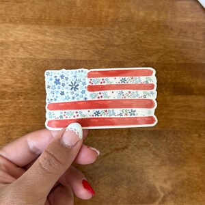 Op de afbeelding: Een rechthoekige sticker met een gestileerd Amerikaans vlagontwerp. Het blauwe kanton en de strepen zijn gevuld met een delicaat patroon van kleine blauwe, rode en witte bloemen en groene blaadjes op een witte achtergrond.
