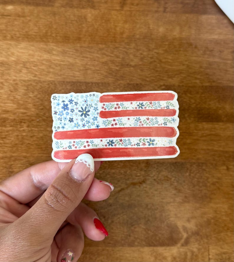 Op de afbeelding: Een rechthoekige sticker met een gestileerd Amerikaans vlagontwerp. Het blauwe kanton en de strepen zijn gevuld met een delicaat patroon van kleine blauwe, rode en witte bloemen en groene blaadjes op een witte achtergrond.