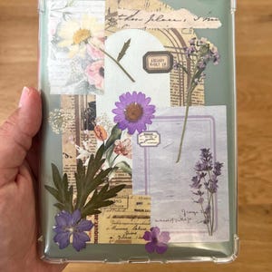 May include: Clear phone case with a collage of dried flowers, vintage paper, and the text "LIBRARY VAULT 19." The design includes purple, pink, and yellow flowers, with green leaves and a light green background.